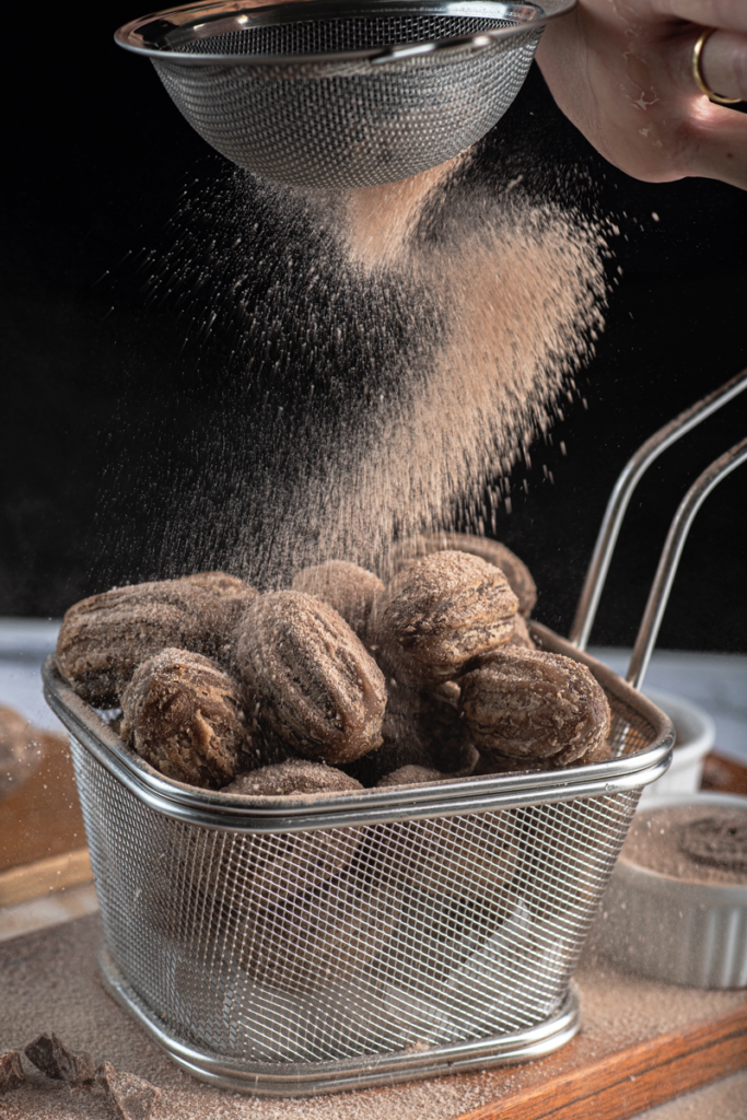 Mini churros com recheio de chocolate em uma cesta metálica com acucar caindo por cima.